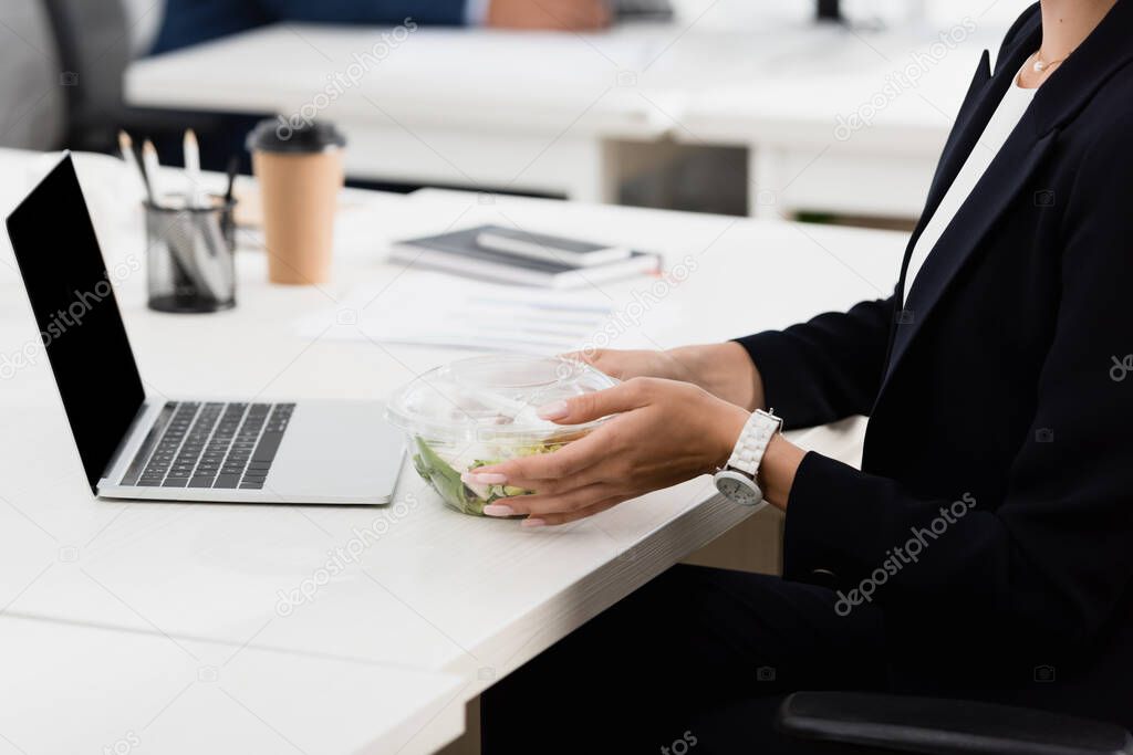 Cropped view of businesswoman holding plastic bowl with meal, while sitting at workplace on blurred background