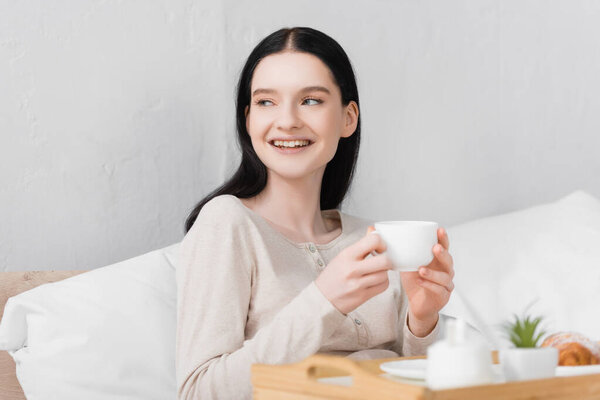 cheerful woman with vitiligo holding cup of tea near breakfast on tray and blurred foreground