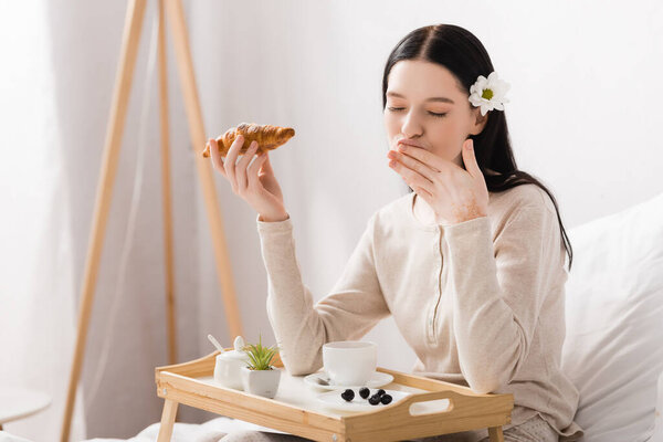 young brunette woman with vitiligo and closed eyes holding croissant near breakfast tray 
