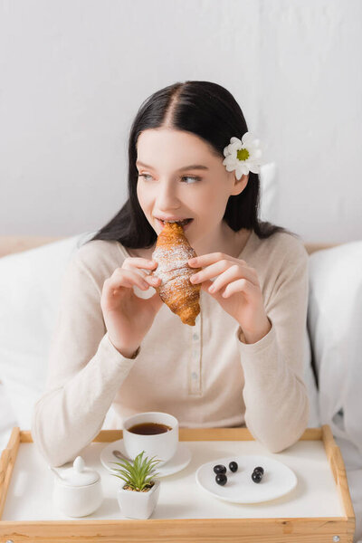 young brunette woman with vitiligo eating croissant near breakfast tray with cup of tea
