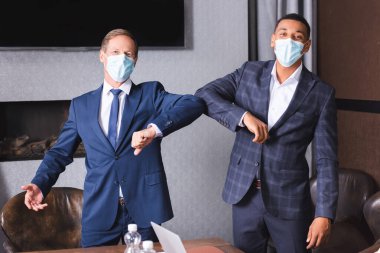 Tıp maskeli çok kültürlü işadamları toplantı odasında dirsekleriyle birbirlerini selamlarken kameraya bakıyorlar.