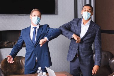 Multicultural businessmen in medical masks looking at camera while greeting each other with elbows in meeting room clipart