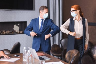 Redhead businesswoman and businessman in medical masks greeting each other with elbows in boardroom clipart