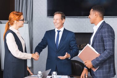 Smiling businesspeople shaking hands with each other while standing near african american colleague in boardroom clipart