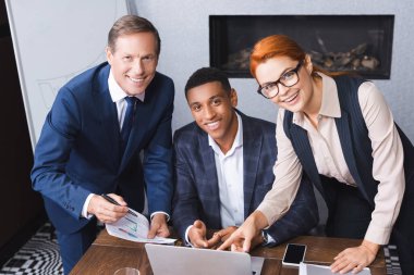Happy multicultural businesspeople looking at camera near workplace with digital devices clipart