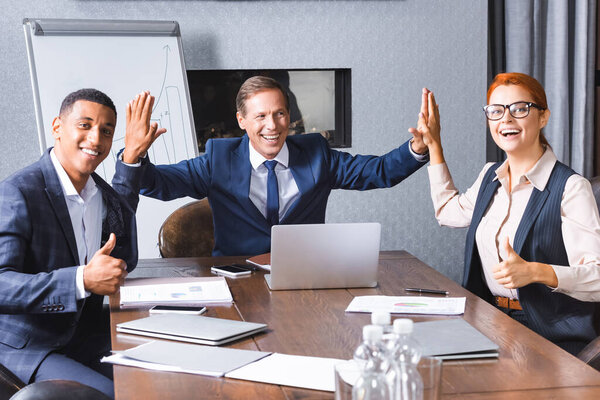 Happy businessman giving high five to multicultural colleagues with thumbs up at workplace in meeting room