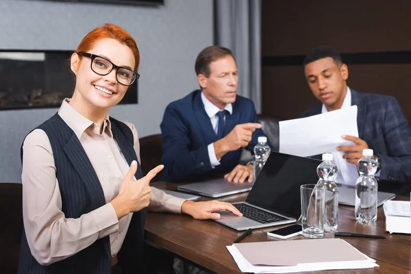 Smiling Businesswoman Looking Camera While Pointing Finger Colleagues Discussing Background — Stock Photo, Image