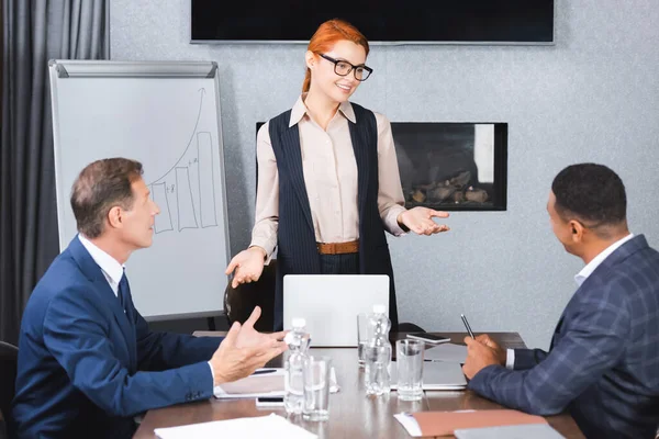 Smiling Redhead Executive Gesturing While Talking Multicultural Colleagues Sitting Workplace — Stock Photo, Image