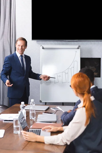 Smiling Executive Pointing Pen Graph Flipchart Blurred Multicultural Colleagues Foreground — Stock Photo, Image