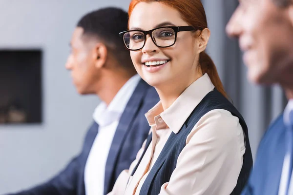 Smiling Redhead Businesswoman Eyeglasses Looking Camera Blurred African American Colleague — Stock Photo, Image