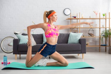 Spor salonundaki halterlerin ve spor şişelerinin yanında spor paspası üzerinde uzanan genç sporcunun yan görüntüsü.
