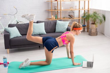 Genç sarışın sporcu laptopun yanında spor paspası, dambıl ve spor şişesi eğitimi alıyor. 
