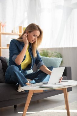 Bacak bacak üstüne atmış genç bir kadın bulanık arka plandaki sehpada dizüstü bilgisayara bakıyor.
