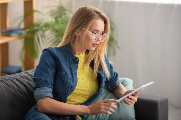 Fiatal Szőke Szemüveges Nézi Digitális Tabletta Kanapén Homályos Háttér — Stock Fotó