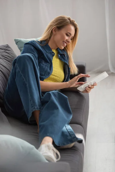 躺在沙发上的金发女人在朦胧的前景中微笑着使用数码平板电脑 — 图库照片