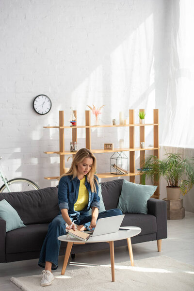 Full length of young blonde woman using laptop while sitting on couch near coffee table at home