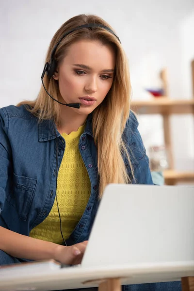 Young blonde woman in headset looking at laptop at home on blurred background