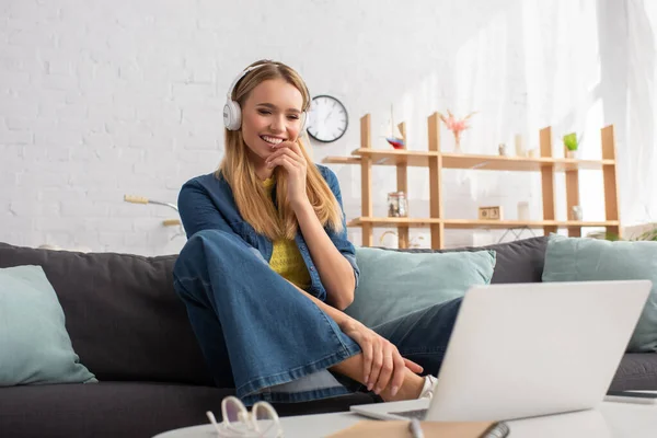 幸せなブロンド女性でヘッドフォン見ますノートパソコンで座っている間にソファに家 — ストック写真
