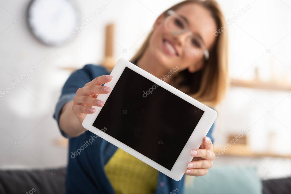 Happy blonde woman looking at camera while showing digital tablet on blurred background