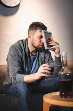 depresif bir adam viski bardağını tutarken yüzünü fotoğraf çerçevesiyle kapatıyor.