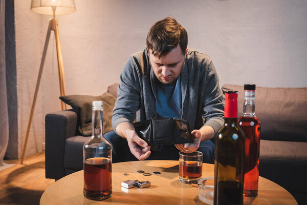 depressed man holding empty wallet near bottles with alcohol drinks at home