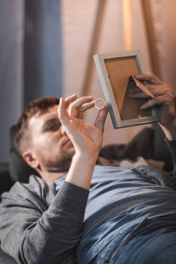Depresyonda, alkol bağımlısı bir adam elinde fotoğraf çerçevesi ve alyans tutarken kanepede yatıyor.