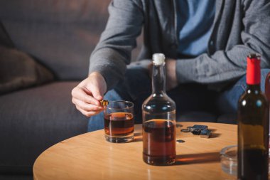Kırpılmış bir görüntü, cam ve viski şişesinin yanında alyans tutan bir adam, bulanık arka plan.