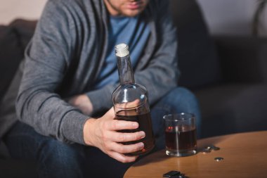 partial view of lonely man holding bottle of whiskey near glass, blurred background clipart
