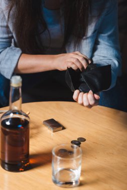 cropped view of alcohol-addicted woman holding empty wallet near bottle of whiskey on blurred foreground clipart