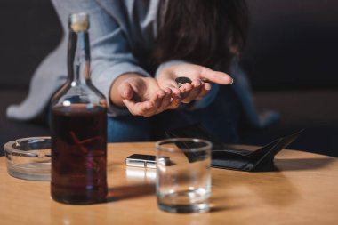 Viskinin yanında bozuk para tutan alkolik bir kadının kısmi görüntüsü ve masanın üzerinde boş bir cüzdan, bulanık arka plan.
