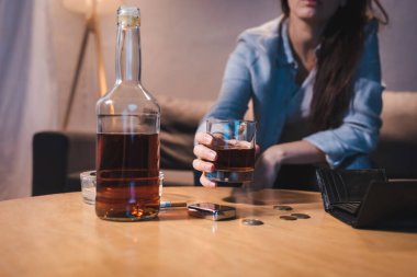 Alkol bağımlısı bir kadının viski bardağını şişenin yanında tutması, bozuk paralar ve boş cüzdan, bulanık arka plan.