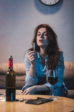 Elinde sigara tutan sarhoş bir kadın kırmızı şarap ve boş bir cüzdanın yanında oturuyor.