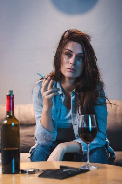 Elinde sigara olan sarhoş bir kadın kırmızı şarabın yanında oturuyor ve ön planda boş bir cüzdan var.  