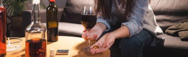 cropped view of woman holding wedding ring and glass of red wine near bottles on blurred foreground, banner clipart