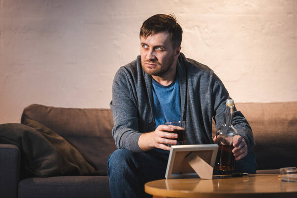 depressed man holding glass and bottle of whiskey near photo frame on table