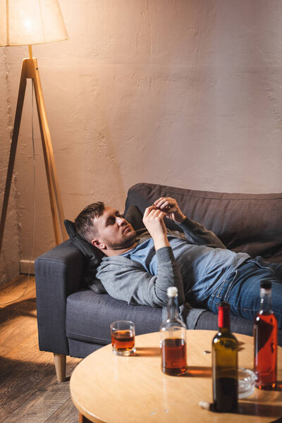 frustrated man lying on sofa near bottles with alcohol drinks on table at home