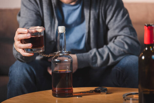 cropped view of alcoholic, handcuffed to bottle, holding glass of whiskey, blurred background