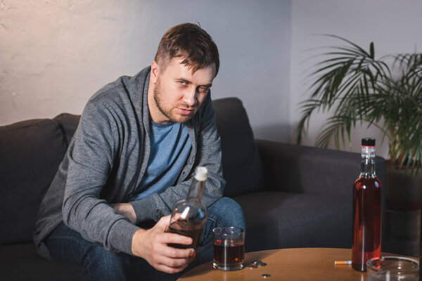 drunk man looking at camera while holding bottle of whiskey at home