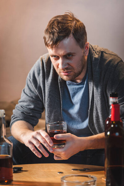 drunk, frustrated man holding glass of whiskey near bottles of alcohol on blurred foreground