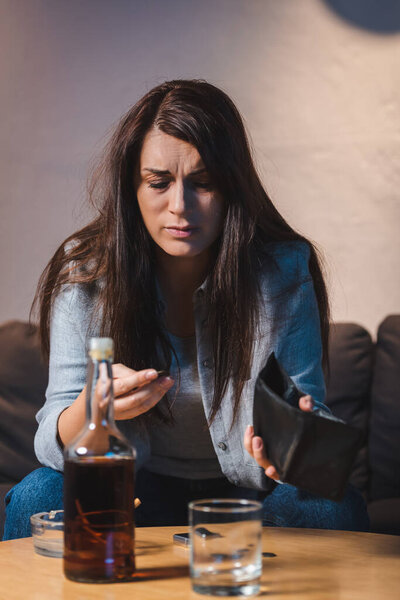 drunk woman holding empty wallet near glass and bottle of whiskey on blurred foreground