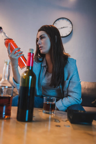 addicted woman holding bottle of cognac near alcohol drinks on blurred foreground