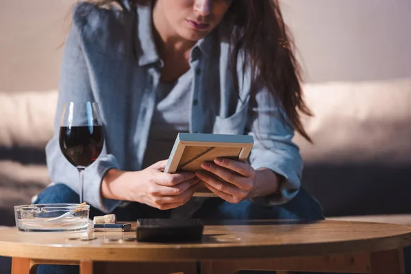 cropped view of depressed woman holding photo frame near glass of red wine, blurred background