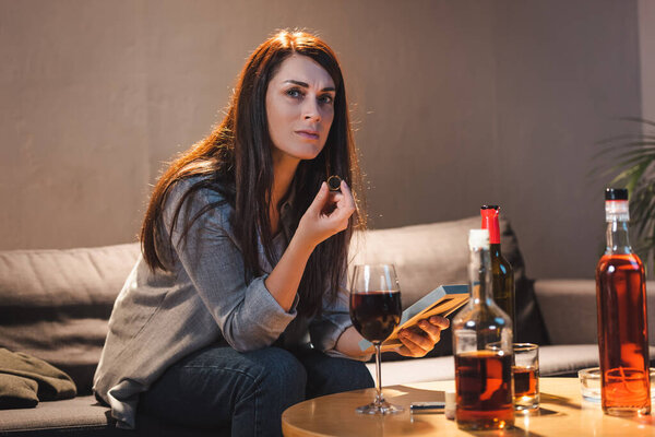 frustrated woman looking at camera while holding wedding ring and photo frame near alcohol drinks on table