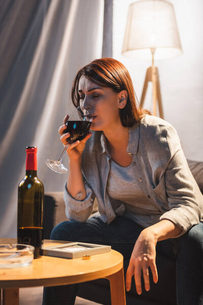 frustrated woman crying and drinking wine while sitting near photo frame and wedding ring on table 