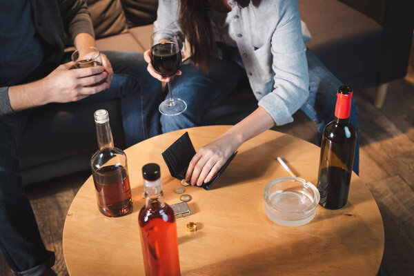 cropped view of couple holding glasses of alcohol near bottles, coins and empty wallet on table