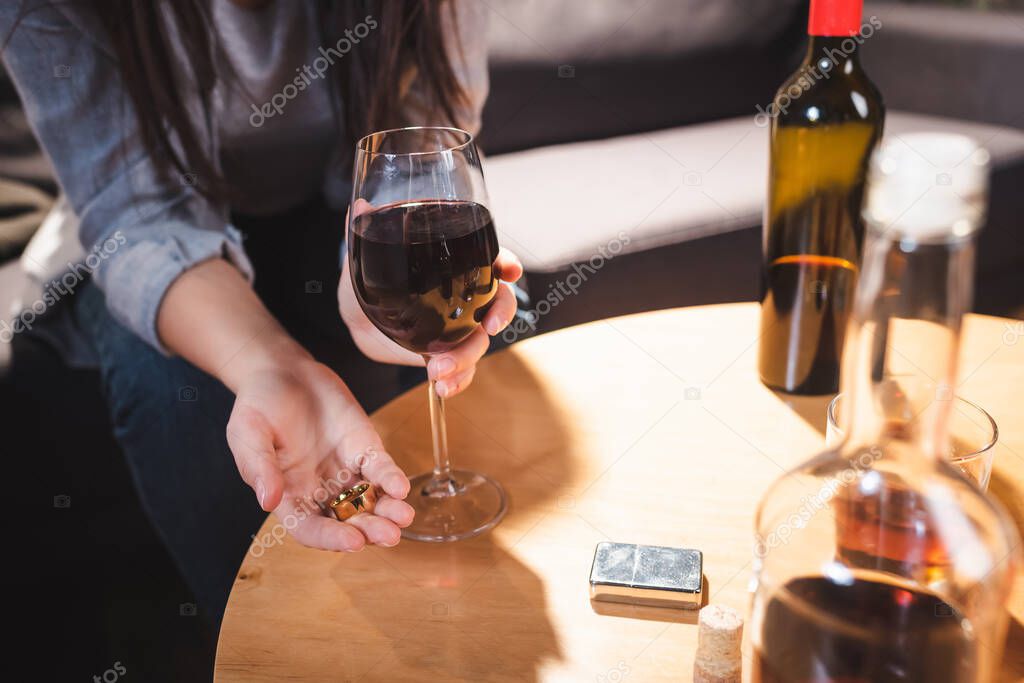 cropped view of alcoholic woman holding wedding rings and glass of wine near bottles on blurred foreground    