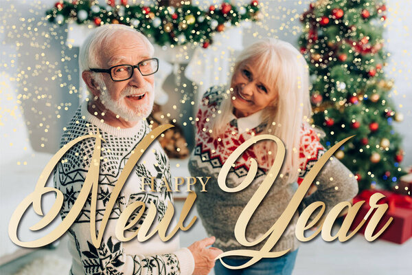 cheerful senior man and woman looking at camera near happy new year lettering and decorations on blurred background 
