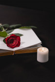 rote Rose auf heiliger Bibel neben Kerze auf schwarzem Hintergrund, Beerdigungskonzept