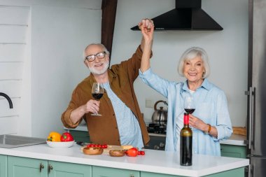 happy senior couple with wine glasses looking at camera while having fun near table with food in kitchen clipart