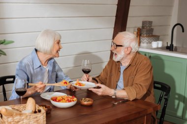 Masada kocasına salata servisi yaparken spatulayla gülen mutlu yaşlı eş ve bulanık arka planda akşam yemeği.